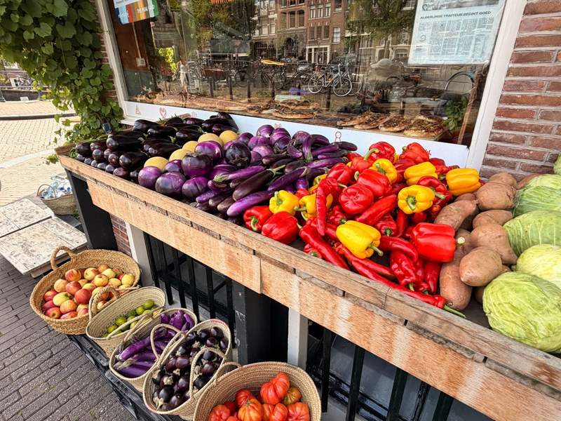 A colorful outdoor produce display in Amsterdam features purple eggplants, red and yellow peppers, cabbage, sweet potatoes, apples and tomatoes arranged outside a neighborhood shop.