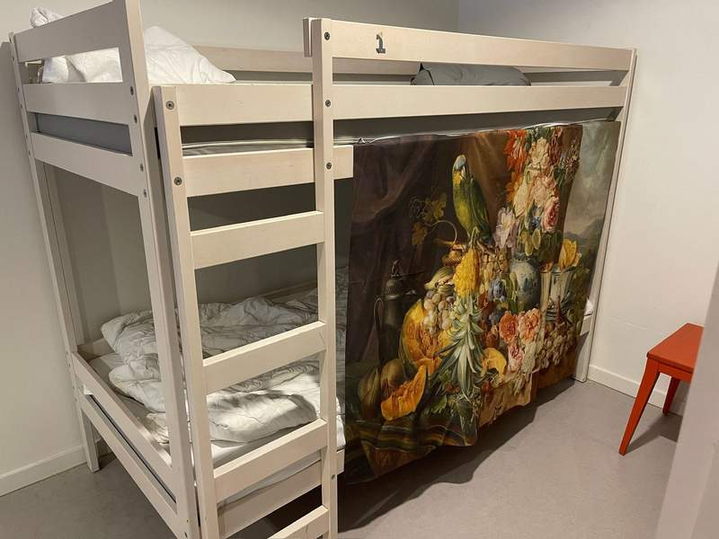 A white wooden bunk bed in a Bruges hostel dorm room features simple white bedding, a ladder and a colorful privacy curtain draped across the lower bunk.
