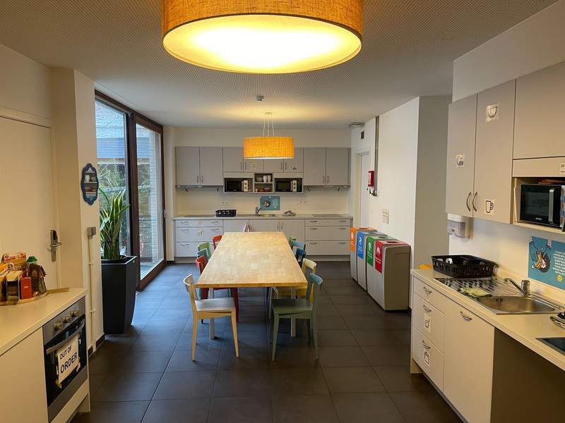 A bright communal hostel kitchen in Bruges, Belgium, features a long dining table, colorful chairs, shared appliances, sinks and cabinets for guest use.