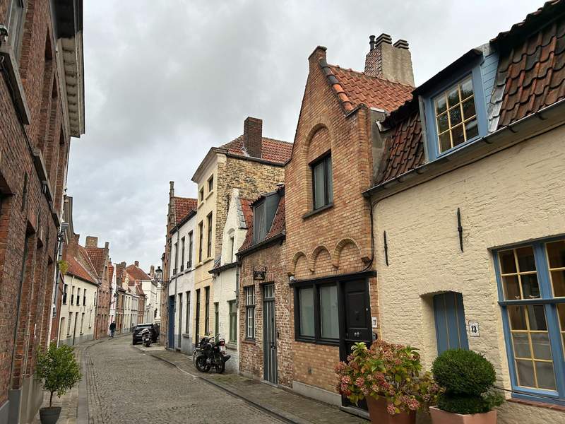 A narrow cobblestone street in Bruges, Belgium, winds past historic brick and stucco homes with red-tiled roofs, colorful windows and potted plants.