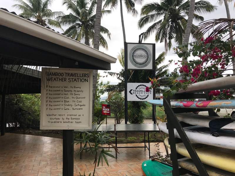 Outdoor common area at Bamboo Travellers hostel in Nadi, Fiji, surrounded by palm trees with surfboards, tropical flowers and a playful weather station sign.
