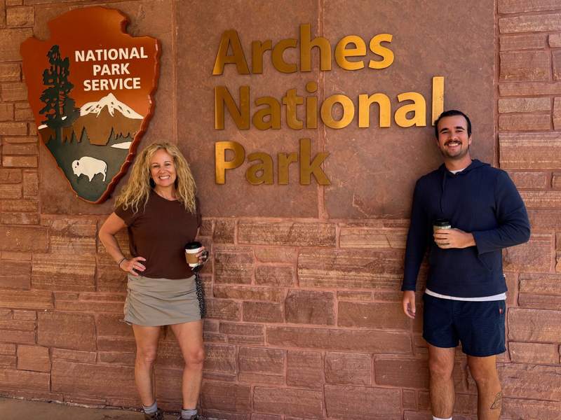 Mother and son holding coffee cups in front of Arches National Park sign.