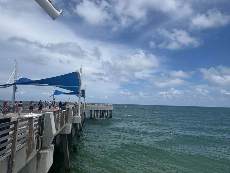 People walking along the Pompano Beach Fisher Family Pier with blue shade sails overlooking the ocean