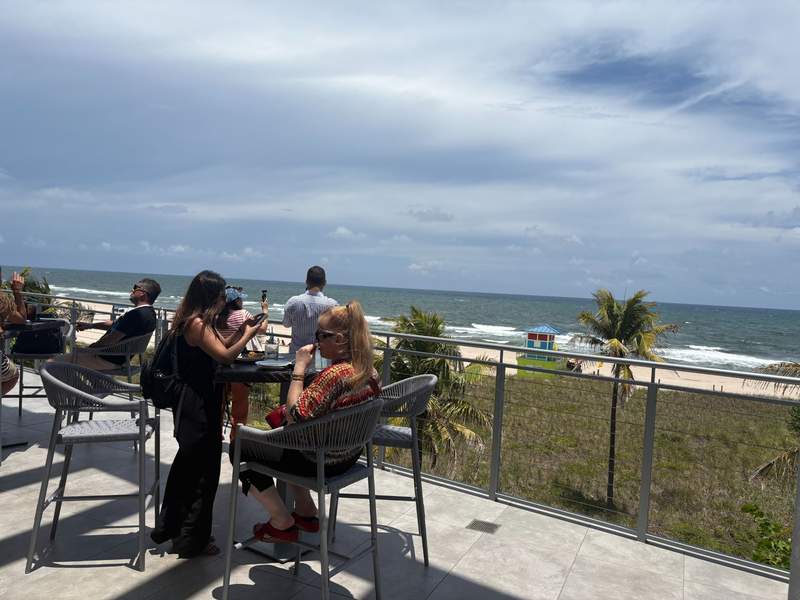 People relaxing on a rooftop terrace at Pompano Pier and Oceanic Restaurant overlooking the ocean beach and palm trees