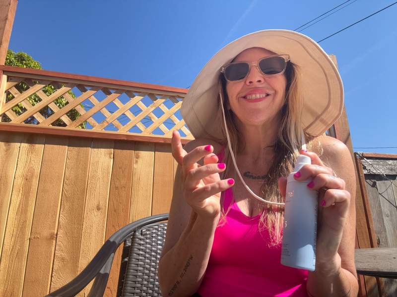 Woman applying sunscreen while wearing sunglasses and a large floppy UPF sun hat.