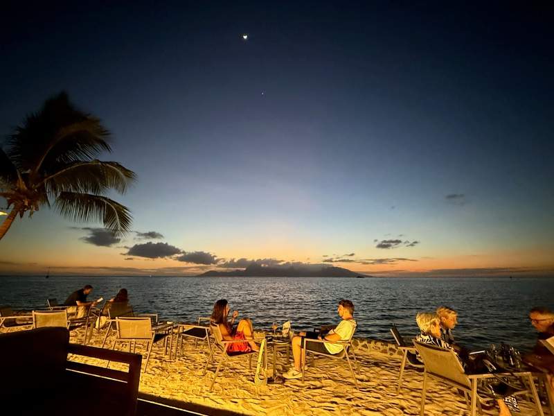 Hotel guests sit around at small tables on the beach enjoying cocktails at sunset.