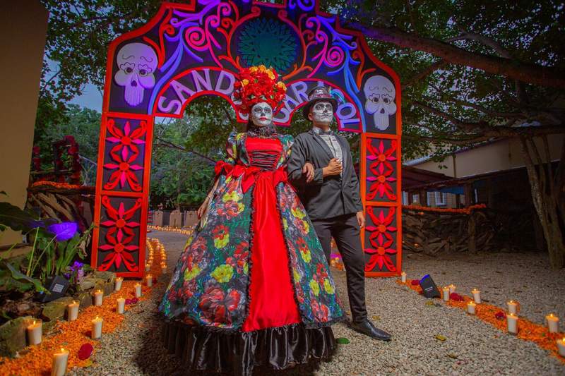 Catrina couple in elaborate Dia de los Muertos costumes beneath a colorful arch at Sandos Caracol Nature Resort & Water Park in Mexico.