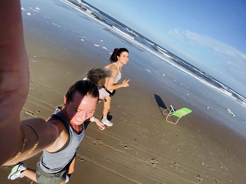 Wide angle beach selfie of smiling woman with two women running behind her on wet sand near ocean green folding chair and blue sky