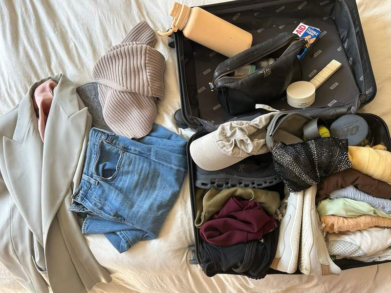 Overhead view of open black suitcase on bed packed with running shoes cap folded clothes toiletry bag water bottle bandages and sequined bag beside blazer sweater and denim shorts