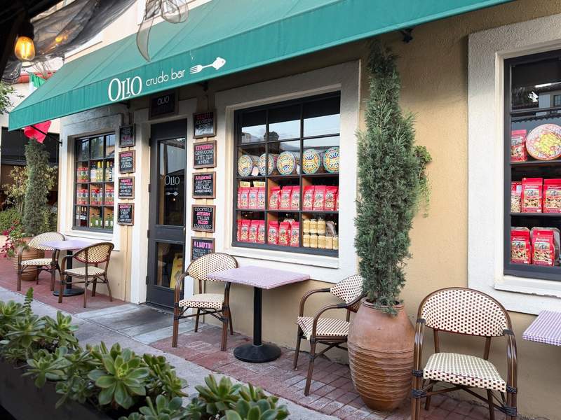 exterior of OLO Crudo Bar in downtown Santa Barbara with a green awning, outdoor café tables and chairs, potted plants and Italian food products displayed in the windows.