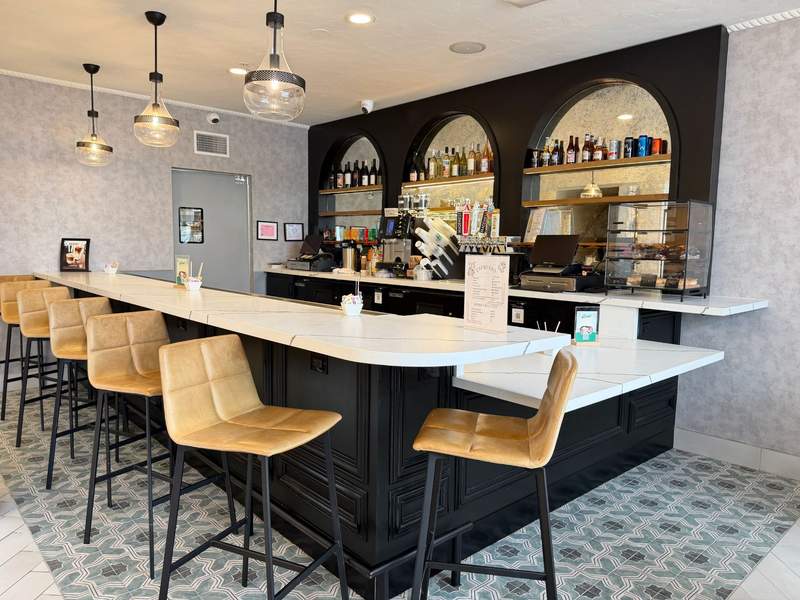 Brisa Café at Hotel Virginia with a long white counter, tan bar stools, coffee equipment, pastry display and bottles of wine, beer and spirits arranged on arched shelves behind the bar.