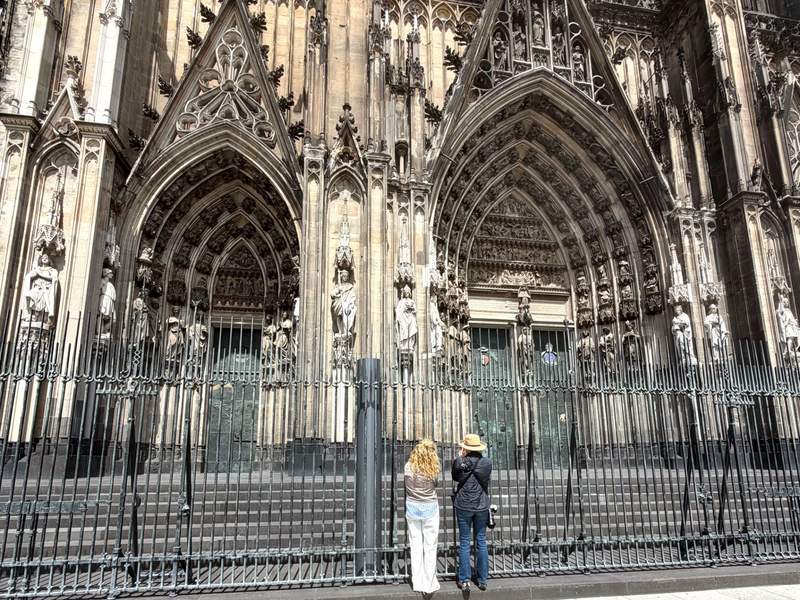 Two women taking a picture behind a fence of an ornate set of church doors with large archways.