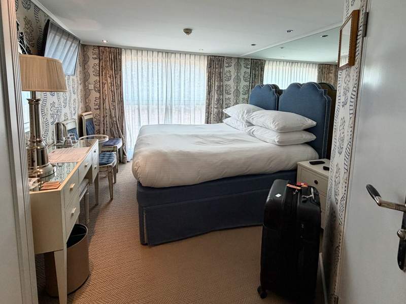 King bed in river cruise stateroom with ornate art deco wallpapers, dresser, chairs, large windows.