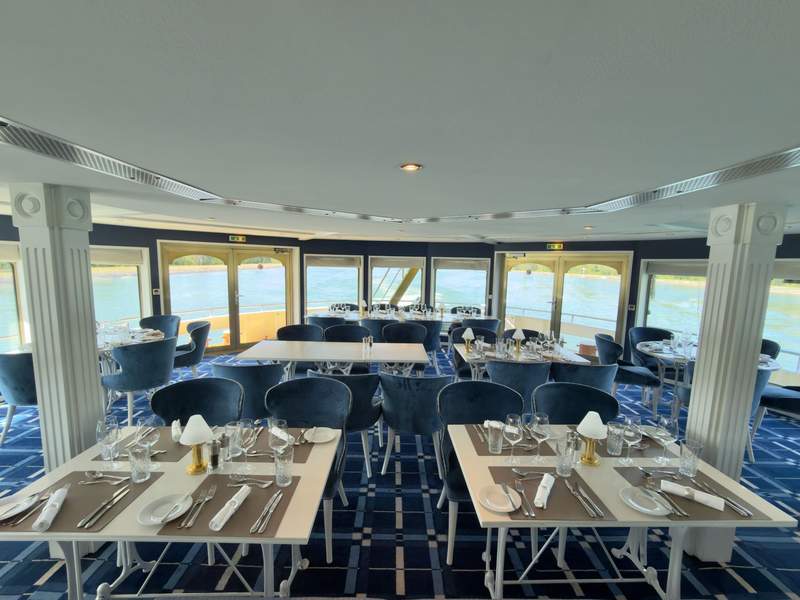 Cruise ship dining hall with blue chairs, blue carpet, white tables and pillars and floor to ceiling windows.