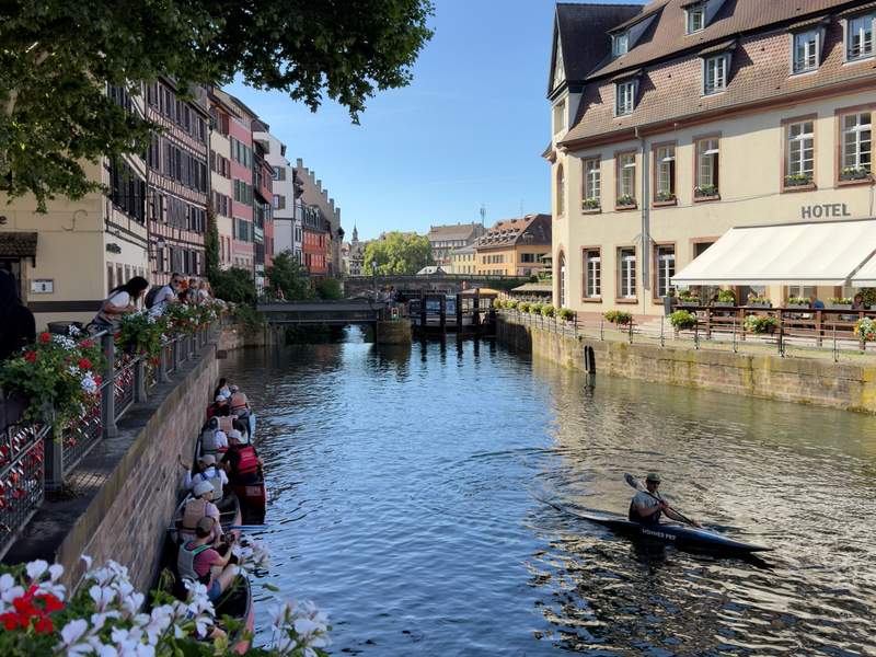 Man paddling a small skiff or kayak in the Ill river flanked by a hotel and colorful buildings and flowers.