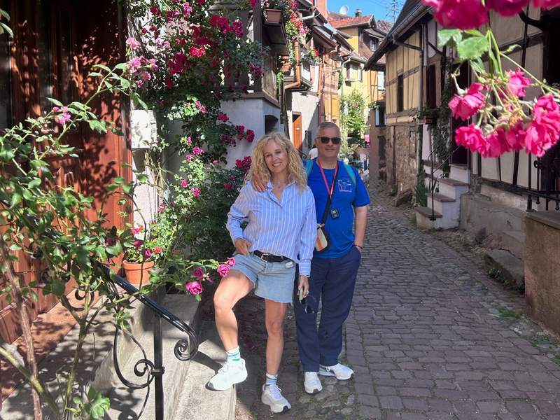 Couple smiling in a colorful archway with pink and red flowers and green vines.