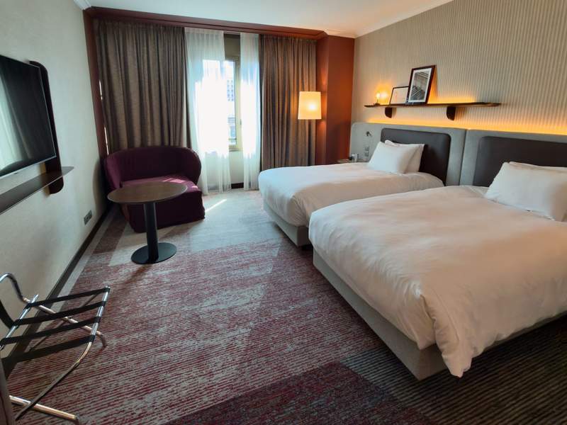 Spacious Hilton Milan guest room with two beds, a large wall-mounted TV, upholstered chair, small table and a window providing natural light.