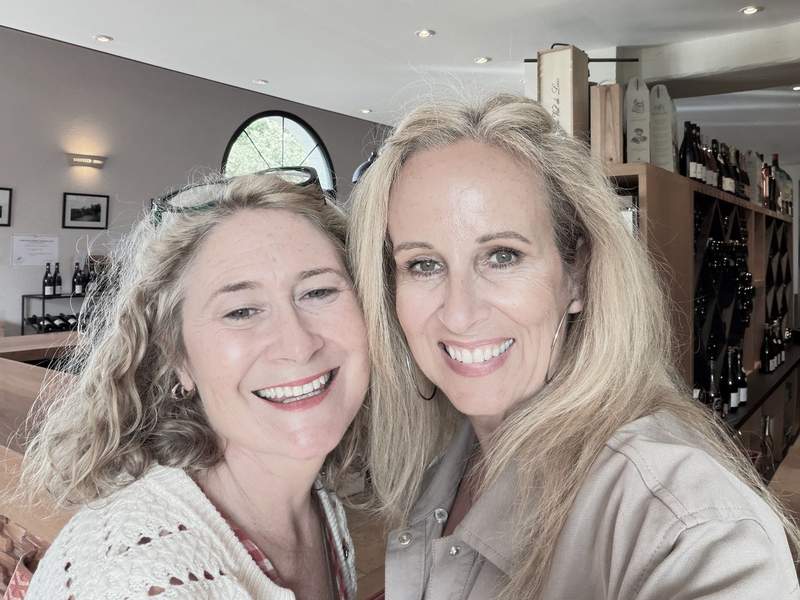 Two smiling women posing cheek to cheek in a bright wine tasting room with wine bottles on shelves behind them