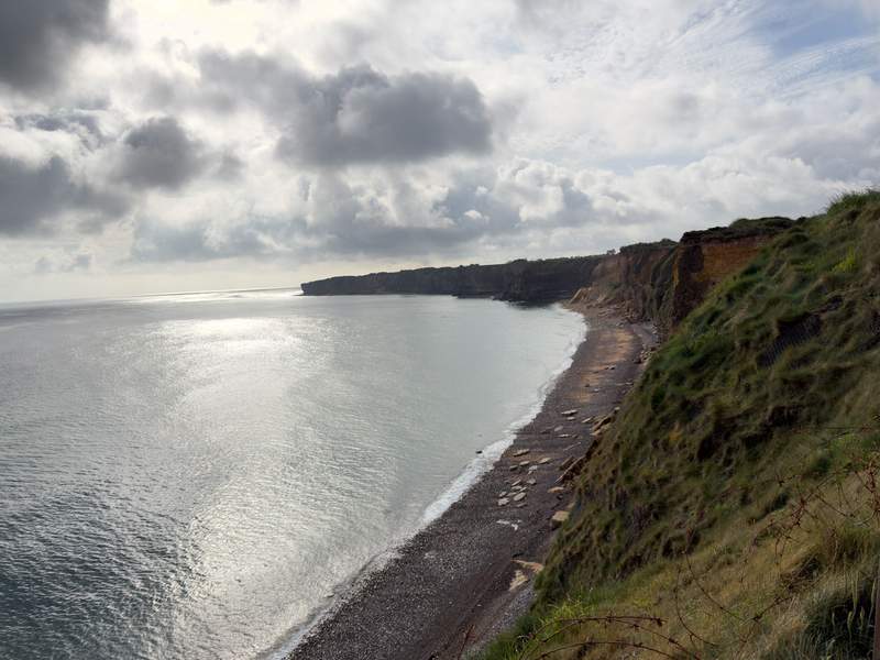 View along steep grassy cliffs and a narrow pebble beach curving toward a headland on the Normandy coast with sunlight on the water