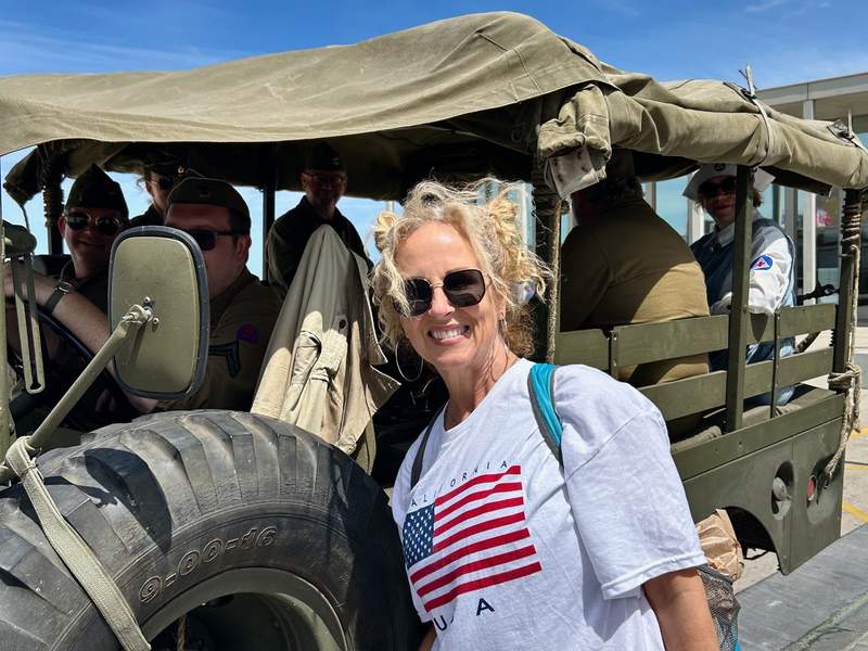 Smiling woman in a California USA flag shirt beside an olive green WWII military truck filled with reenactors in period uniforms