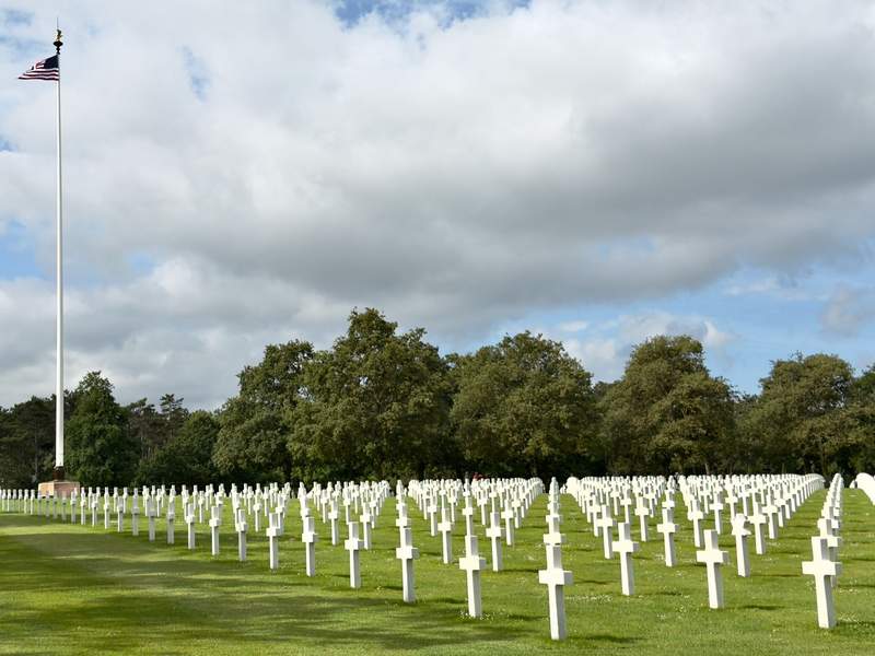 Rows of white marble crosses on green lawn at the Normandy American Cemetery with an American flag flying on a tall flagpole.