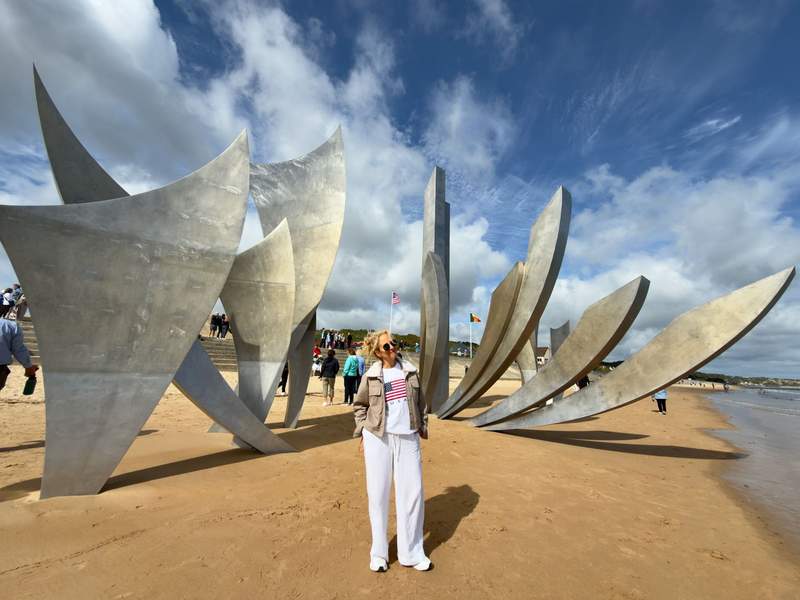 Woman in a white USA flag shirt standing in front of the tall stainless steel Les Braves sculpture on the sand at Omaha Beach under a blue sky