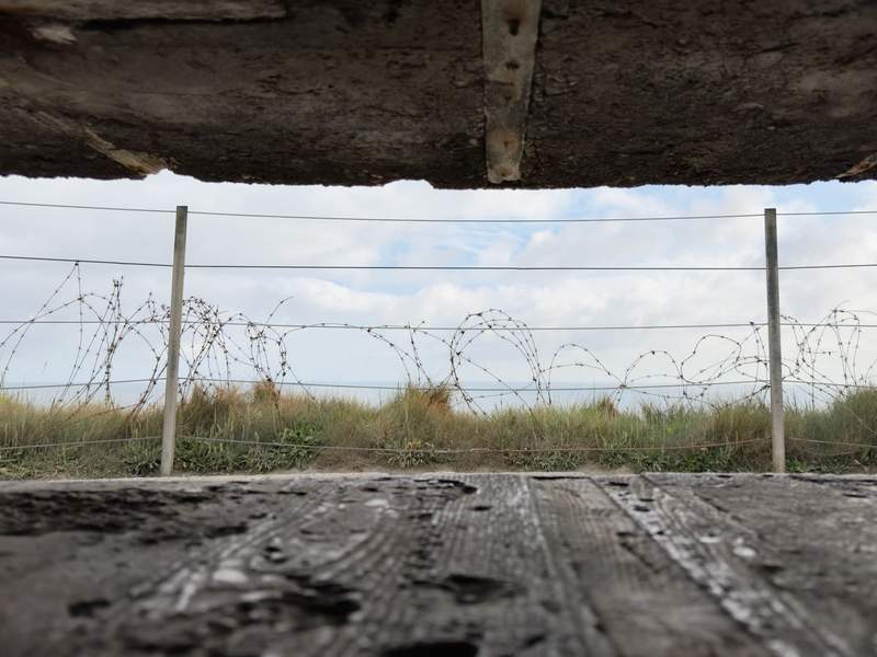 View through a narrow concrete bunker opening showing coiled barbed wire and grass with the sea in the distance.