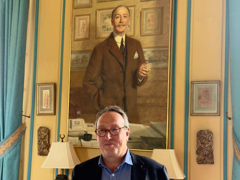 Man in round glasses and a navy blazer standing beneath a large oil portrait of a mustached gentleman in a yellow and turquoise room