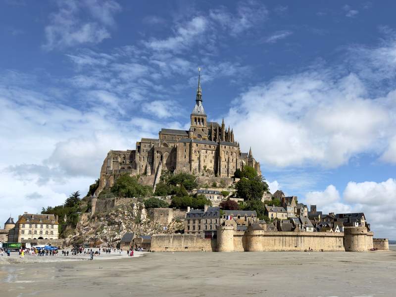 Mont Saint Michel abbey rising above medieval ramparts and village houses on the tidal flats under a bright blue sky
