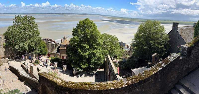Panoramic view from the stone ramparts of Mont Saint Michel over trees, rooftops and visitors toward wide sandy tidal flats and blue sky