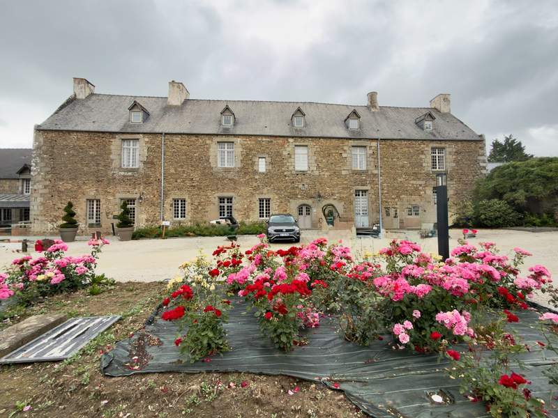 Long granite manor house with slate roof and dormer windows behind a bed of pink, red and yellow roses under gray skies