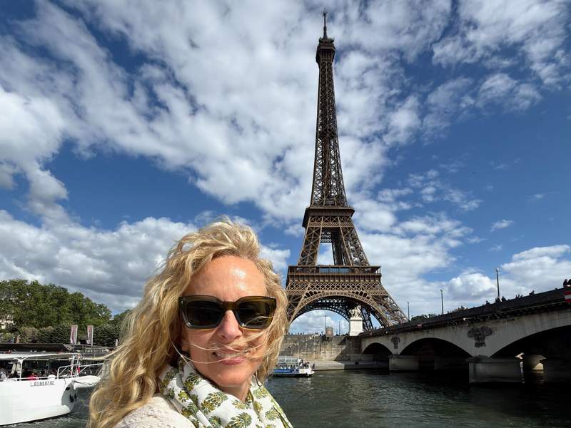 Woman with windblown curly blonde hair and sunglasses taking a selfie on the Seine with the Eiffel Tower and Pont d'Iéna behind her