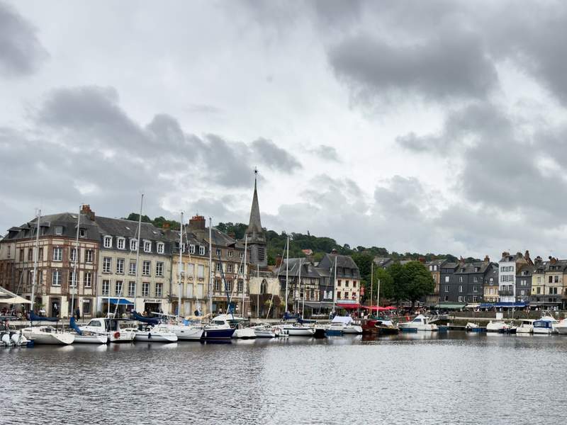 Sailboats moored in Honfleur's old harbor in front of tall slate and timber buildings and a church spire under gray clouds