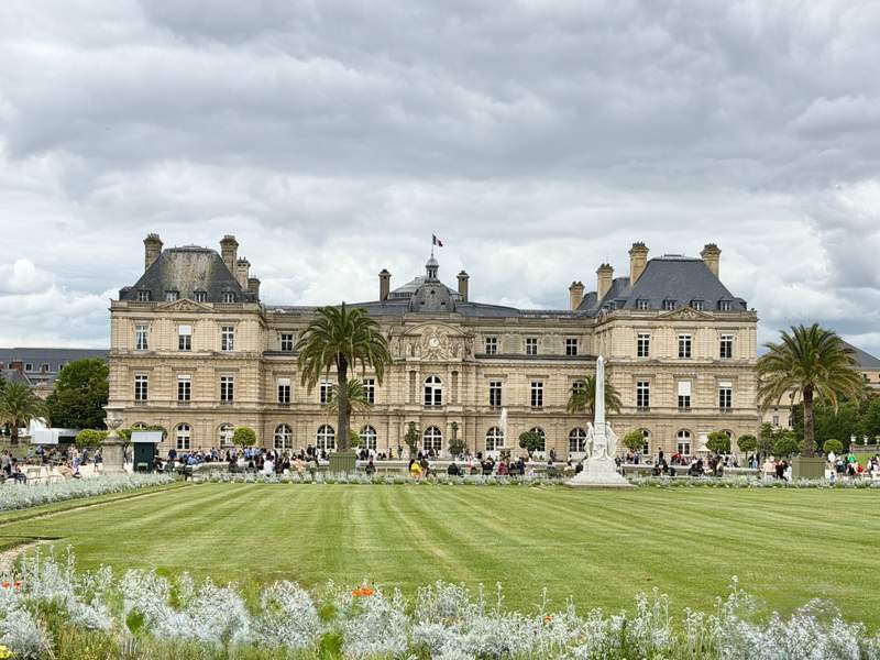Luxembourg Palace in Paris behind a striped green lawn, palm trees and white flowers under cloudy skies