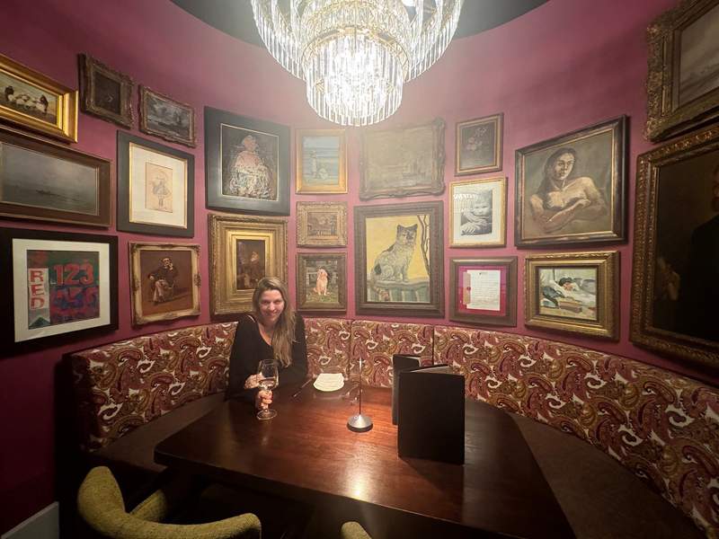 The writer is pictured seated in a circular restaurant booth surrounded by a gallery wall of framed paintings.