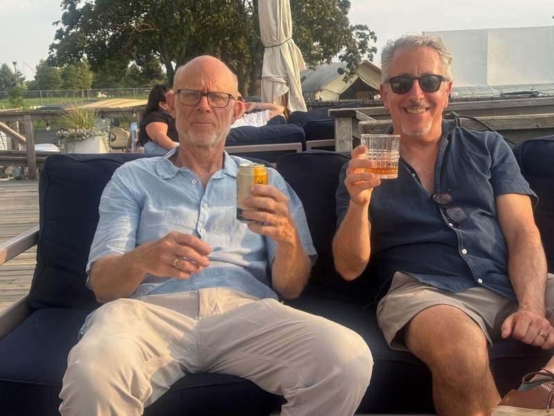 Two men sitting outdoors on a dark couch, with the man on the left wearing glasses and holding a canned drink, and the man on the right wearing sunglasses and raising a glass of liquor for a toast