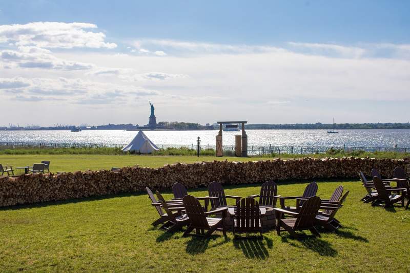 A grassy lawn featuring a circle of wooden Adirondack chairs around a fire pit, a low wall made of stacked firewood, a white tent nearby, and a scenic waterfront view in the background with the Statue of Liberty visible across the water.