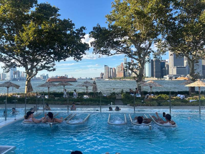 People relaxing in a pool with built-in loungers, overlooking a waterfront with umbrellas, trees, and a distant city skyline.