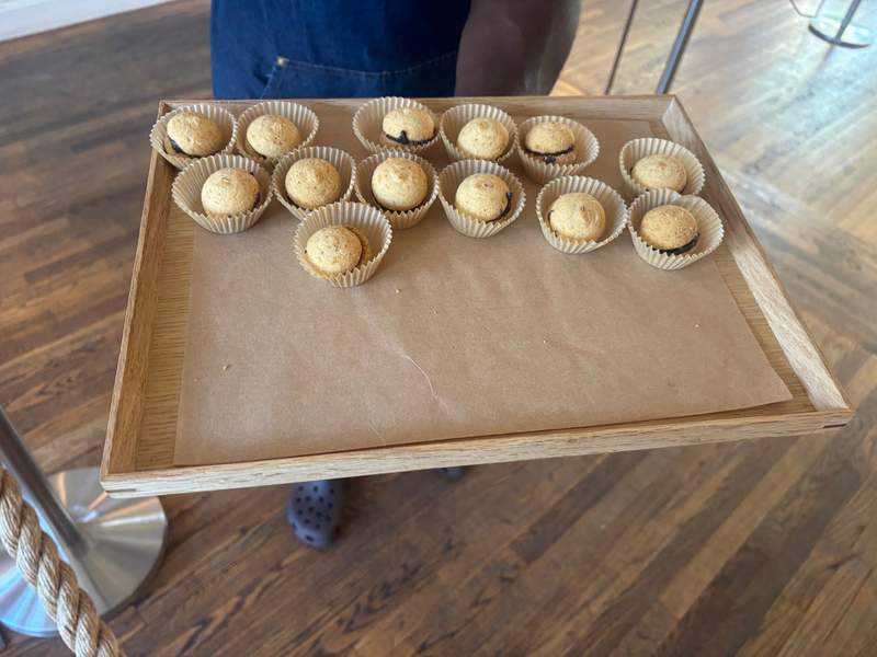 A wooden tray lined with brown paper holds a selection of small, round baked treats in individual paper liners, being held by someone standing on a wooden floor.