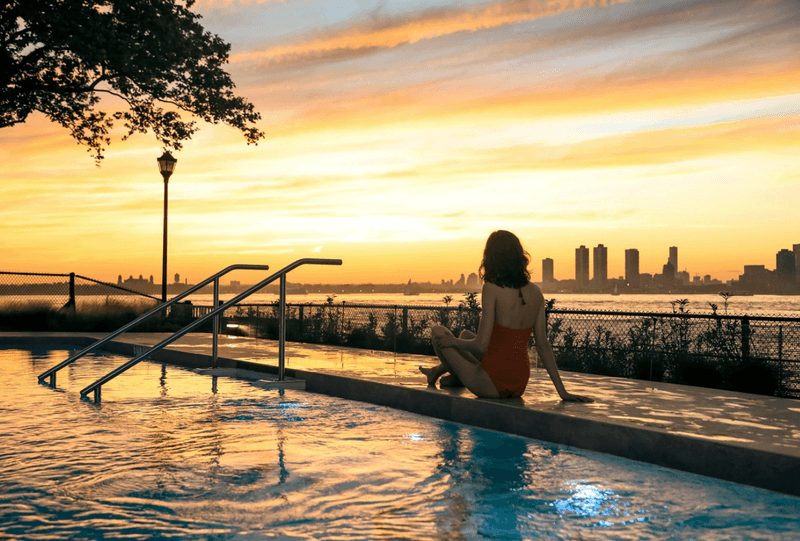A person sits on the edge of an outdoor swimming pool at sunset, looking out toward a waterfront city skyline reflecting golden light across the sky and water.