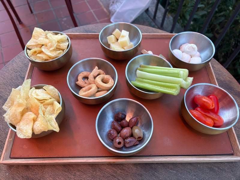 A tray arranged with small metal bowls containing various snacks, including potato chips, cubed cheese, small round crackers, celery stalks, olives, cherry tomatoes, and round white snacks.