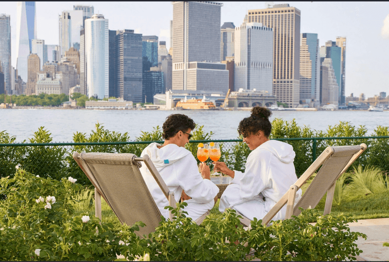 Two people wearing white bathrobes sit in lounge chairs outdoors among green bushes, facing a body of water while clinking glasses filled with orange cocktails
