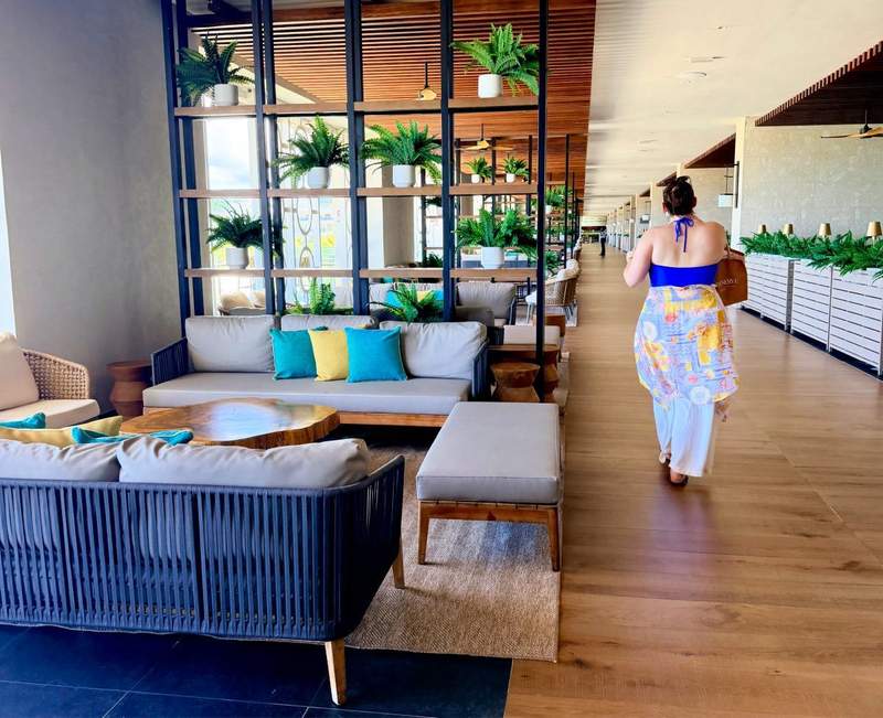 Open-air walkway along Restaurant Row at Princess Grand Jamaica featuring stylish lounge seating, tropical plants, modern décor and a guest strolling toward the oceanfront restaurants overlooking the Caribbean Sea.
