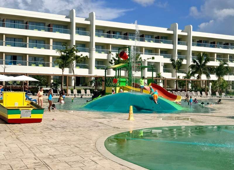 Children splash and play in the Princess Grand Jamaica aquatic park featuring colorful water slides, fountains, a shallow kids' pool, and family-friendly accommodations in the background.