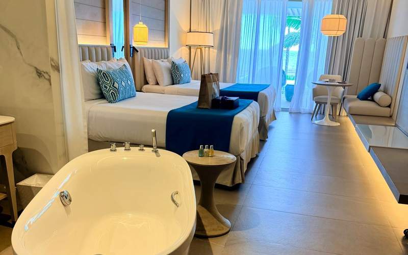 Modern guest room at Princess Grand Jamaica featuring two queen beds, a soaking tub, contemporary furnishings, and a private balcony with tropical resort views.