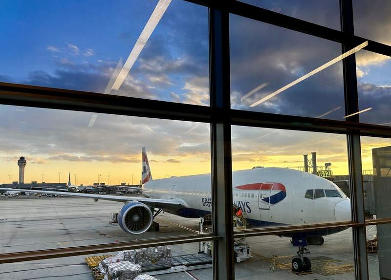 British Airways aircraft parked at a gate at London Heathrow Airport, seen through terminal windows against a golden evening sky