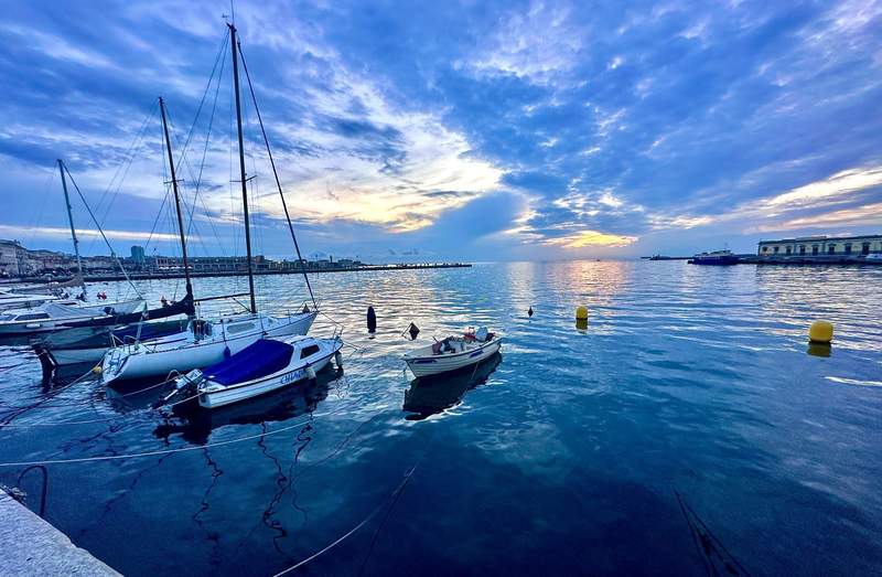 Sailboats and small boats along the Trieste waterfront at dusk, with calm water reflecting a dramatic blue and golden sky