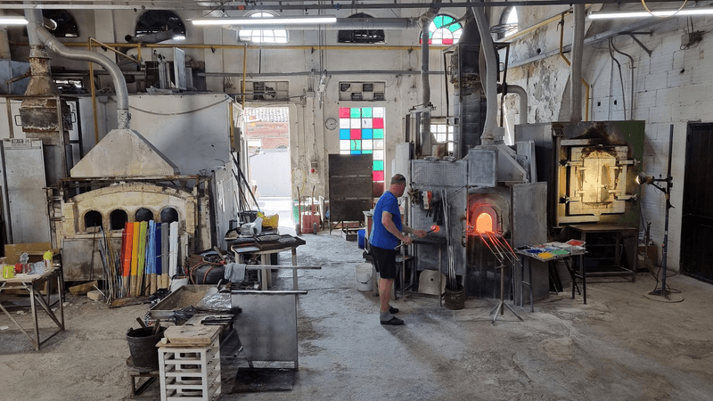 An industrial warehouse with a glass-blowing furnace, colored glass rods, and an artisan demonstrating glassblowing. 