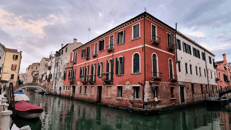 An old corner building in Venice with red paint and green shutters, surrounded on two sides by dark green canals. 