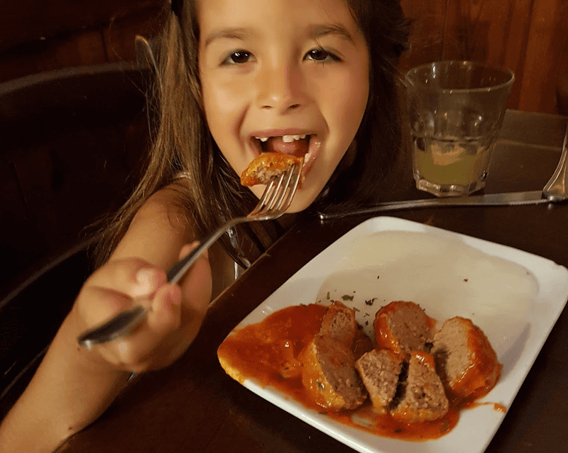 A little girl with a missing front tooth and a satisfied look on her face eats a plate of meatballs. 